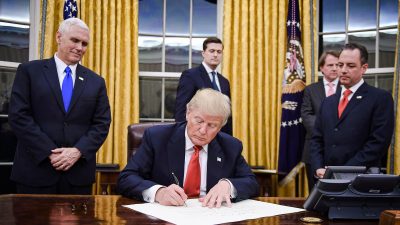 Президент Дональд Трамп подписывает указ, а вице-президент Майк Пенс и глава администрации Райнс Прибус наблюдают за этим в Белом доме 20 января 2017 года. (Jim Watson/AFP via Getty Images)