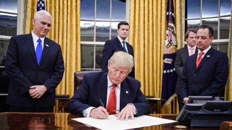 Президент Дональд Трамп подписывает указ, а вице-президент Майк Пенс и глава администрации Райнс Прибус наблюдают за этим в Белом доме 20 января 2017 года. (Jim Watson/AFP via Getty Images)