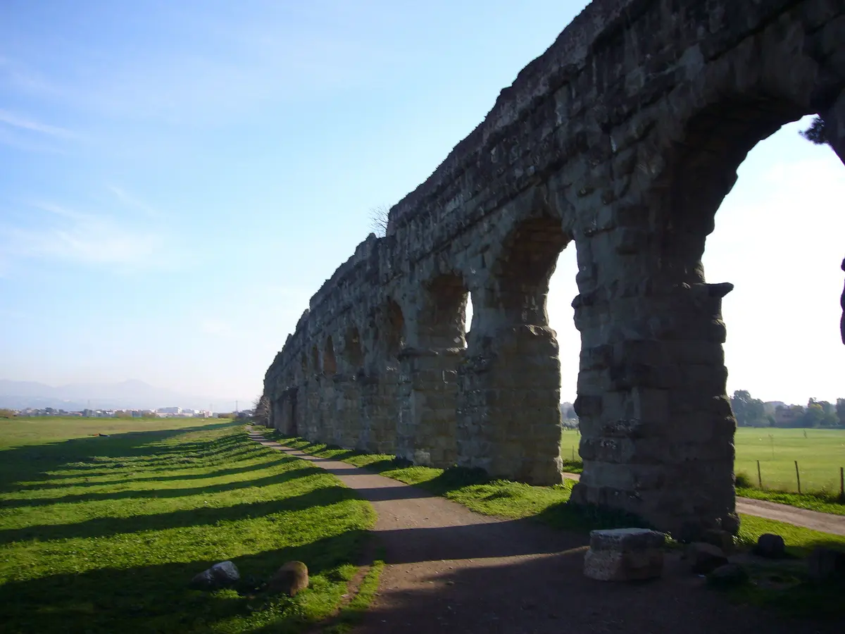 Древнеримский каменный акведук с арками на фоне зеленого поля и голубого неба.