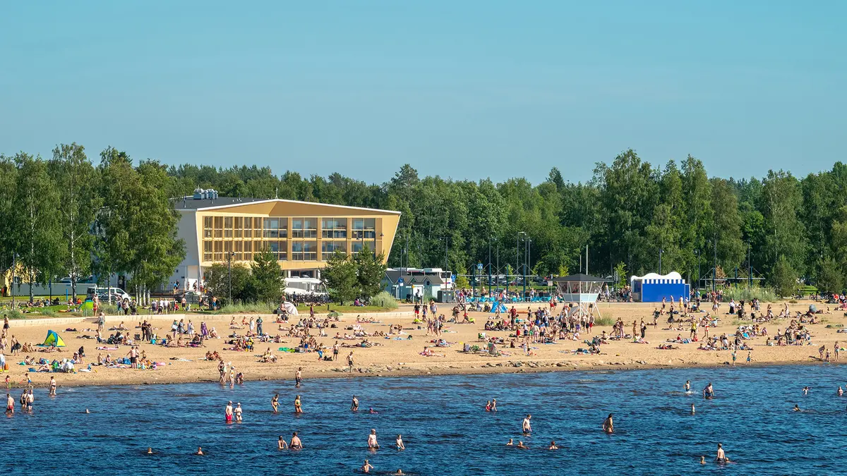 Люди отдыхают на песчаном пляже Хиетаниеми в Хельсинки на фоне желтого здания павильона в солнечный день.