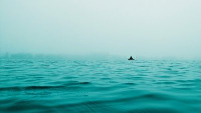 Вдали виднеется крошечный силуэт человека в маленькой лодке на фоне бескрайнего моря с небольшими волнами в бирюзово-голубых тонах.
