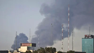 Столбы густого дыма над горизонтом Тегерана на фоне антенн и городских зданий во время военного конфликта.