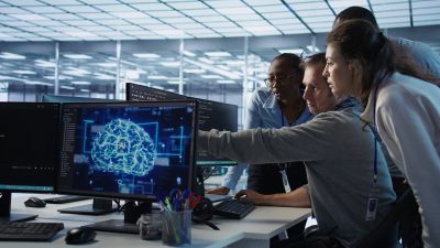 Команда IT-специалистов обсуждает код и визуальную схему ИИ на мониторах.